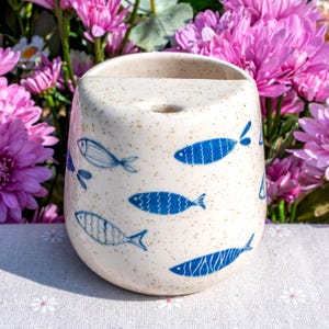 Könnte beinhalten: Eine gesprenkelte Keramikvase mit geschwungener Form und einer oberen Öffnung. Die Vase ist mit blauen Fischillustrationen verziert. Der Hintergrund zeigt rosa Blüten und erzeugt einen lebendigen Kontrast. Die Vase dient wahrscheinlich zum Halten von Blumen oder anderen dekorativen Gegenständen.