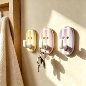 Op de afbeelding: Drie ovale wandhaken met verticale strepen in geel, roze en paars. Elke haak heeft een witte gebogen arm en twee kleine ronde schroefgaten. Een set zilverkleurige metalen sleutels hangt aan de middelste roze haak aan de muur bevestigd.