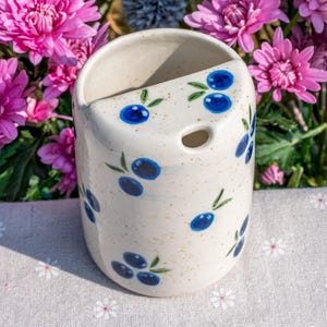 May include: A ceramic wall pocket with a cream-colored base, speckled with brown flecks. The pocket is adorned with hand-painted blue olives and green leaves. It features a hole for hanging and is set against a backdrop of pink flowers.