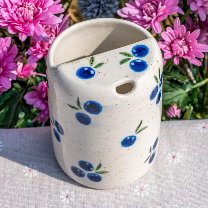 May include: A ceramic wall pocket with a cream-colored base, speckled with brown flecks. The pocket is adorned with hand-painted blue olives and green leaves. It features a hole for hanging and is set against a backdrop of pink flowers.
