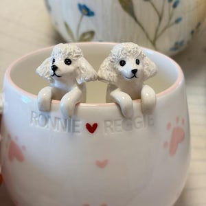 May include: A white ceramic mug featuring two sculpted white poodle figures peering over the rim. The mug is decorated with small pink paw prints and the embossed text "RONNIE &hearts; REGGIE" in the center, with a light pink rim around the top edge.