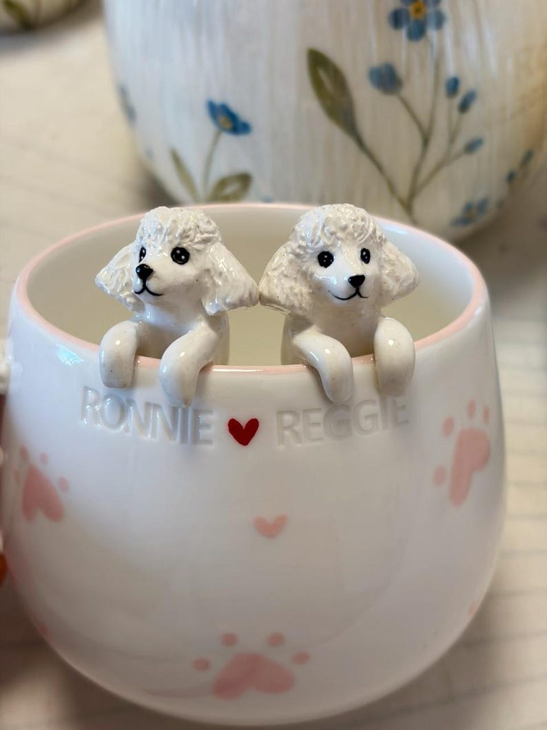 May include: A white ceramic mug featuring two sculpted white poodle figures peering over the rim. The mug is decorated with small pink paw prints and the embossed text "RONNIE &hearts; REGGIE" in the center, with a light pink rim around the top edge.