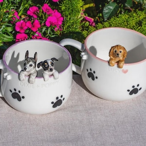 May include: Two white ceramic mugs featuring black paw prints and hand-painted dog figurines peering over the rims. One mug is inscribed with the text DIXIE + POPPY, while the other features the name Lucy next to a small pink heart graphic.