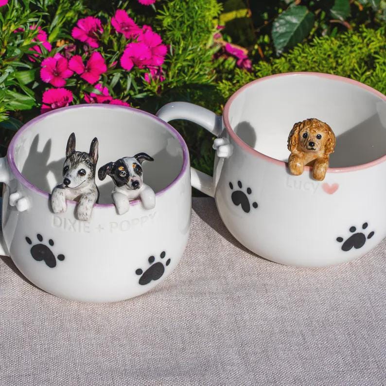 May include: Two white ceramic mugs featuring black paw prints and hand-painted dog figurines peering over the rims. One mug is inscribed with the text DIXIE + POPPY, while the other features the name Lucy next to a small pink heart graphic.