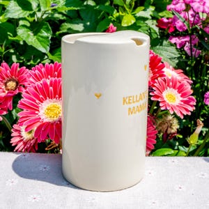May include: A cream-colored ceramic mug with a lid, featuring the text "Kellan's Mama" in gold lettering. The mug has a heart-shaped detail and is set against a backdrop of pink and yellow flowers.