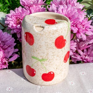 May include: A ceramic, cylindrical container with a speckled, off-white finish. The container is adorned with bright red cherry illustrations and green stems. The background features vibrant pink flowers, suggesting a decorative or kitchenware item.