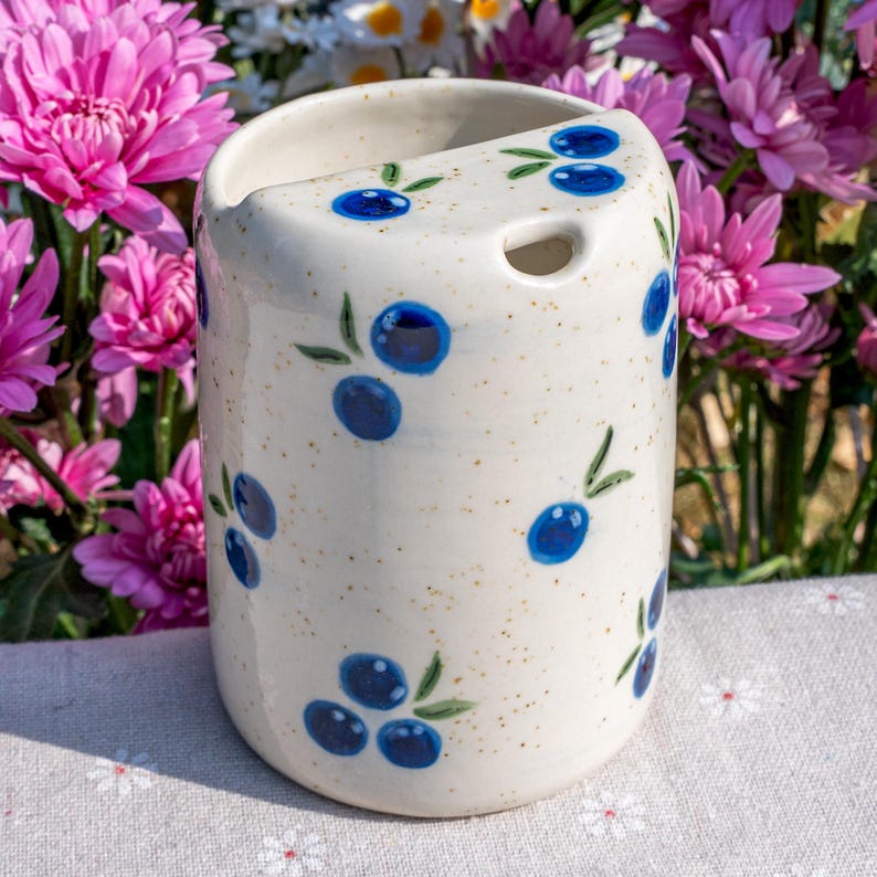May include: A white ceramic toothbrush holder with a speckled finish, adorned with hand-painted blue blueberries and green leaves. The holder has a curved top and a small hole. The design is reminiscent of a rustic, farmhouse style.