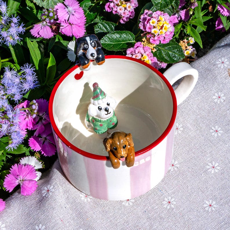 May include: A white ceramic mug with pink vertical stripes and a red rim. The mug is decorated with three small dog figurines. The background features colourful flowers and a patterned cloth.