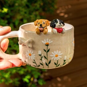 Puede incluir: Una taza de cer&aacute;mica moteada con un patr&oacute;n de margaritas pintado a mano y un hueso esculpido en el asa. Dos peque&ntilde;as figuras de perro tridimensionales, una dorada y otra negra y blanca con un pa&ntilde;uelo rojo de lunares, asoman por el borde.