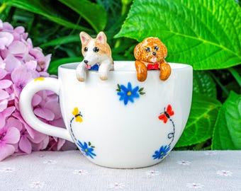 May include: A white ceramic mug featuring hand-painted blue flowers and small butterflies, with two miniature dog figurines perched on the rim. One figurine is a tan and white corgi with a blue bowtie, and the other is a brown poodle with a red bowtie.