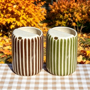 May include: Two cylindrical ceramic containers with a speckled cream-colored top and vertical stripes. One container features dark brown stripes, while the other features olive green stripes. Each piece has a small circular opening near the rim.