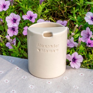 May include: A cream ceramic vase with a rectangular opening, inscribed with "Alexander Mama". The vase is set against a backdrop of purple flowers and green leaves.