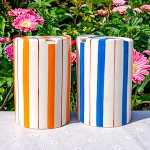 May include: Two cylindrical ceramic containers featuring vertical stripes. One container displays alternating orange and thin brown stripes on a cream background, while the other features alternating blue and thin red stripes on a cream background.
