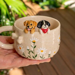 Puede incluir: Una taza de cer&aacute;mica beige con acabado moteado, que presenta un cachorro marr&oacute;n dorado esculpido y un cachorro blanco y negro con un pa&ntilde;uelo rojo de lunares asom&aacute;ndose por el borde. El exterior est&aacute; decorado con margaritas blancas y tallos verdes.