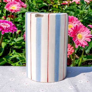 May include: A cylindrical ceramic utensil holder featuring a cream-colored base with vertical light blue stripes and thin, vibrant red pinstripes. The top edge has a small, semi-circular cutout, and the surface has a smooth, glossy finish.