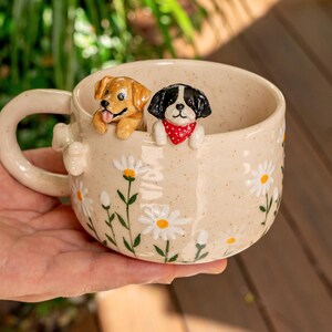 Puede incluir: Una taza de cer&aacute;mica color crema con acabado moteado, decorada con margaritas blancas pintadas con centros amarillos. Dos peque&ntilde;as figuras de perros esculpidas est&aacute;n unidas al borde interior: un cachorro marr&oacute;n dorado y un perro blanco y negro con pa&ntilde;uelo rojo.