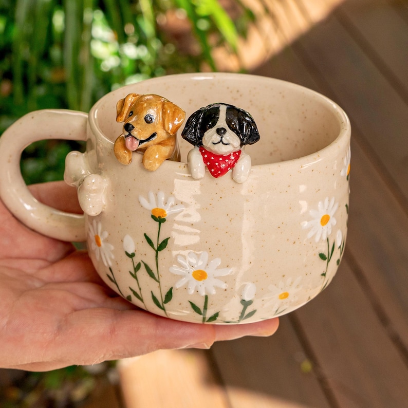 Puede incluir: Una taza de cer&aacute;mica color crema con acabado moteado, decorada con margaritas blancas pintadas con centros amarillos. Dos peque&ntilde;as figuras de perros esculpidas est&aacute;n unidas al borde interior: un cachorro marr&oacute;n dorado y un perro blanco y negro con pa&ntilde;uelo rojo.