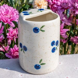 May include: A speckled off-white ceramic toothbrush holder with a curved top and a slot. The holder is decorated with hand-painted blue blueberries and green leaves. The background features pink and yellow flowers.