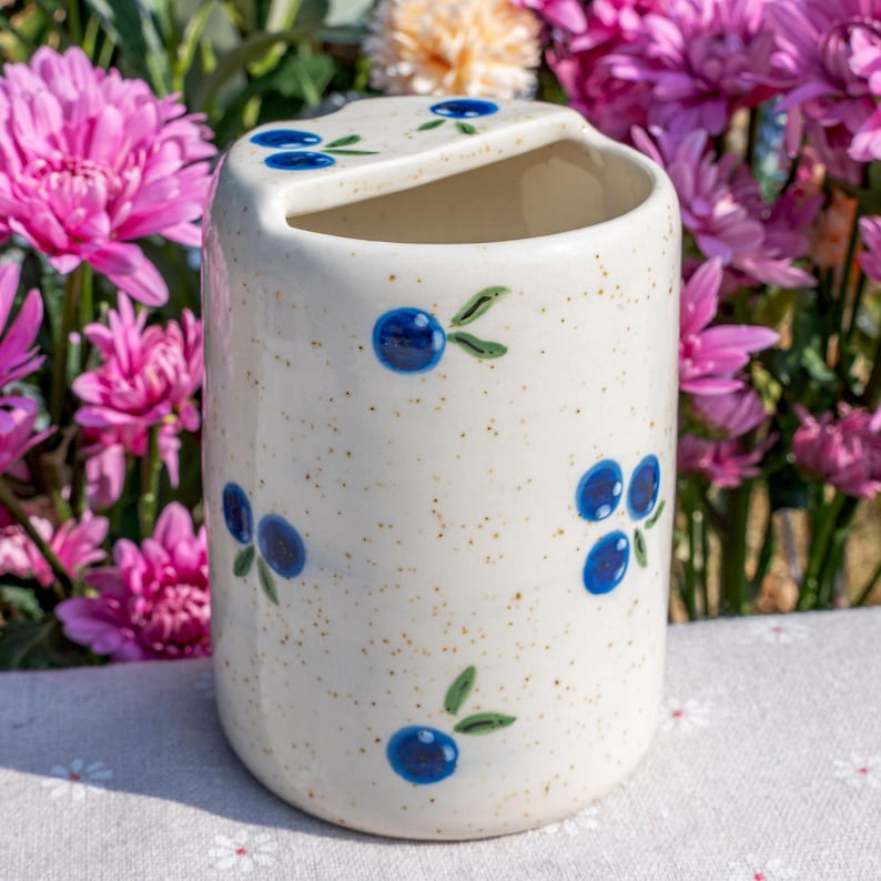 May include: A speckled off-white ceramic toothbrush holder with a curved top and a slot. The holder is decorated with hand-painted blue blueberries and green leaves. The background features pink and yellow flowers.