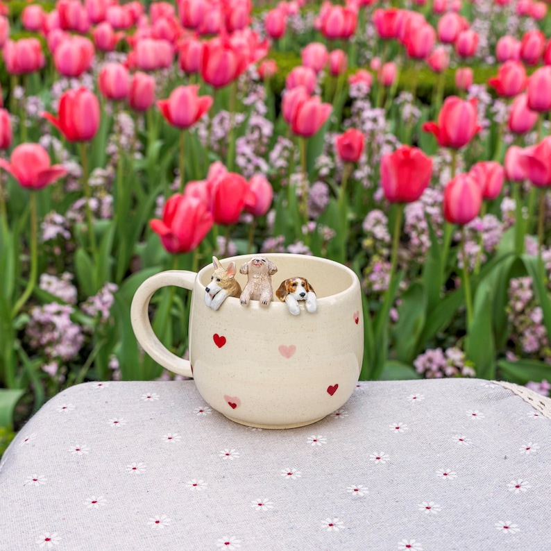 K&ouml;nnte beinhalten: Eine wei&szlig;e Keramiktasse mit roten Herzen bedruckt. Drei kleine Hundefiguren schauen &uuml;ber den Rand der Tasse. Die Tasse steht auf einer wei&szlig;en Tischdecke mit Blumenmuster.