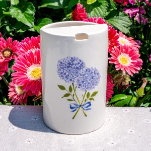 May include: A white ceramic container with a rounded top and a rectangular opening. The container is decorated with a blue hydrangea floral design and a blue ribbon. The background features pink flowers and green foliage.