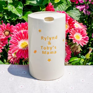 May include: A white ceramic tumbler with the text "Rylind & Toby's Mama" in gold lettering, along with gold star accents. The tumbler has a small opening at the top and is surrounded by pink and yellow flowers.