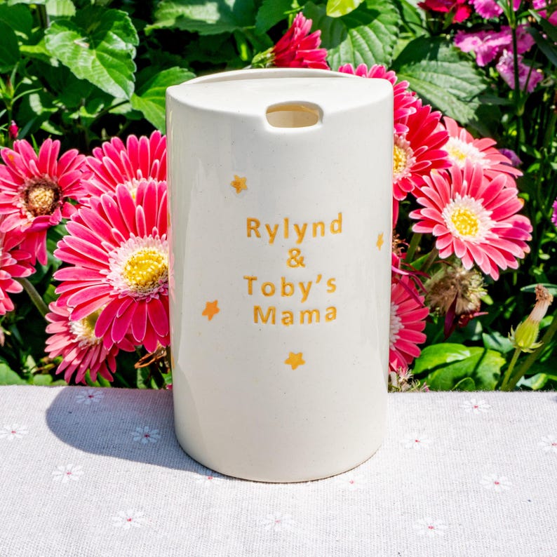 May include: A white ceramic tumbler with the text "Rylind & Toby's Mama" in gold lettering, along with gold star accents. The tumbler has a small opening at the top and is surrounded by pink and yellow flowers.