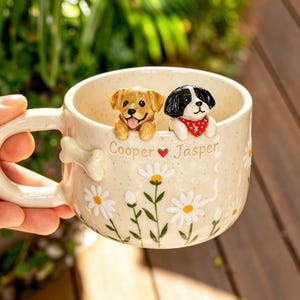 Puede incluir: Una taza de cer&aacute;mica color crema con acabado moteado, que presenta dos figuras de perro esculpidas asom&aacute;ndose por el borde. La taza muestra el texto Cooper, un coraz&oacute;n rojo y Jasper, junto con margaritas blancas pintadas y un asa con forma de hueso.