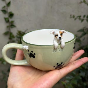 May include: A light green ceramic mug featuring a three-dimensional white and brown puppy figure peering over the rim. The exterior is decorated with black paw prints and the name JUNIE embossed in the center. It has a curved handle.