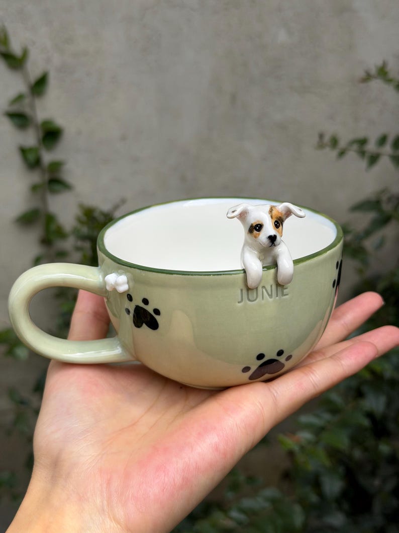 May include: A light green ceramic mug featuring a three-dimensional white and brown puppy figure peering over the rim. The exterior is decorated with black paw prints and the name JUNIE embossed in the center. It has a curved handle.