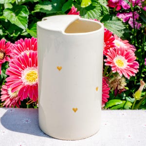 May include: A cream-colored ceramic vase with a unique curved top and small gold heart accents. The vase is cylindrical and stands against a backdrop of vibrant pink flowers with yellow centers, creating a cheerful and decorative aesthetic.