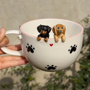 May include: A white ceramic mug with a light pink rim, featuring black paw prints and the text REX ❤️ CECE. Two small, sculpted dog figurines, one black and tan and one golden brown, are attached to the inner rim, appearing to peek over the edge.