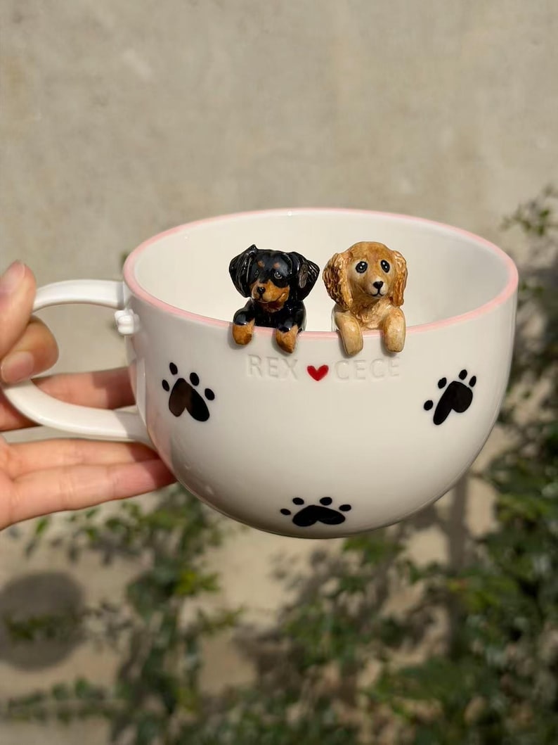 May include: A white ceramic mug with a light pink rim, featuring black paw prints and the text REX ❤️ CECE. Two small, sculpted dog figurines, one black and tan and one golden brown, are attached to the inner rim, appearing to peek over the edge.