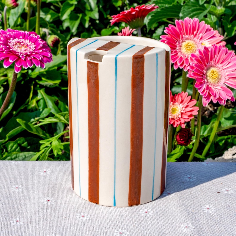 May include: A cylindrical ceramic utensil holder with a cream-colored base featuring vertical brown stripes and thin light blue pinstripes. The top edge has a small semi-circular cutout, and the surface is smooth with a glossy finish.