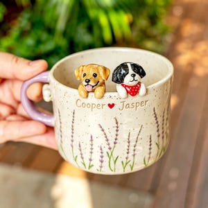 May include: A speckled ceramic mug with a light purple handle, featuring two sculpted dog figurines peering over the rim. The mug is decorated with lavender sprigs and the text Cooper heart Jasper printed in brown on the front surface.
