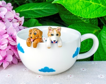 May include: A white ceramic mug featuring blue cloud patterns on the exterior. Two small, hand-painted dog figurines are attached to the inner rim, one brown and one tan and white, both appearing to peek over the edge of the cup.