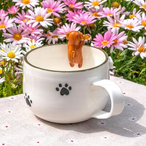 May include: A white ceramic mug featuring a small, three-dimensional brown dog figurine attached to the inner rim. The exterior of the mug is decorated with several black paw print patterns and has a gold-toned rim along the top edge.