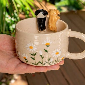 Puede incluir: Una taza de cer&aacute;mica beige con acabado moteado, decorada con margaritas blancas pintadas a mano con centros amarillos. Dos peque&ntilde;as figuras de perro de cer&aacute;mica tridimensionales, una negra con blanco y otra color canela, est&aacute;n en el borde.