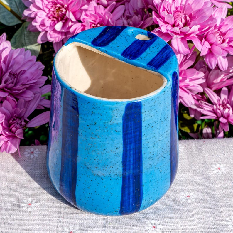 Peut inclure: Un cache-pot mural en c&eacute;ramique avec un &eacute;mail bleu clair mouchet&eacute; et des rayures verticales bleu fonc&eacute;. Le r&eacute;cipient pr&eacute;sente une large ouverture inclin&eacute;e sur le dessus et un petit trou circulaire pour la suspension, avec une finition lisse et brillante.