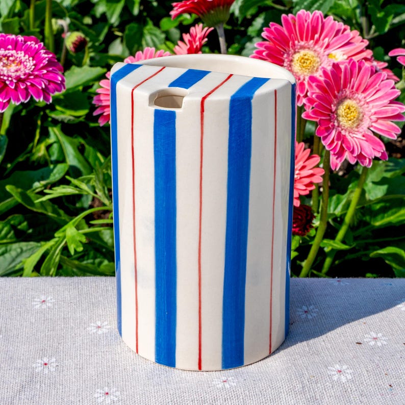 May include: A cylindrical ceramic vessel with a cream-colored base featuring vertical blue and thin red stripes. The top edge is curved, and there is a small circular opening near the upper rim of the object, which has a smooth, glossy finish.