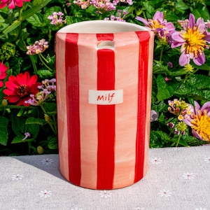May include: A cylindrical ceramic vase featuring vertical red and light pink stripes. A small white rectangular label is centered on the front with the text Milf written in red script. The top opening is circular with a smooth white interior rim.