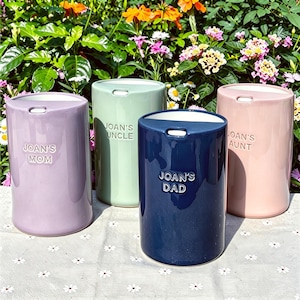 May include: Four cylindrical ceramic containers in lavender, light green, navy blue, and light pink. Each features a white lid and embossed text: JOAN'S MOM, JOAN'S UNCLE, JOAN'S DAD, and JOAN'S AUNT. The containers have a smooth, glossy finish.