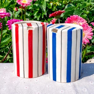 May include: Two cylindrical ceramic containers with open tops. One container features vertical red and thin black stripes on a white background. The second container features vertical blue and thin black stripes on a white background.