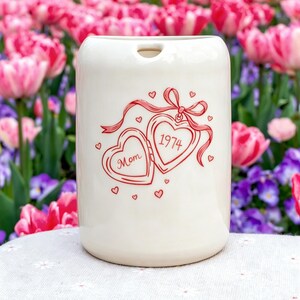 May include: A cylindrical off-white ceramic vessel featuring a red line-art illustration of an open heart-shaped locket tied with a ribbon. The left side of the locket reads "Mom" and the right side reads "1974," surrounded by small red hearts.