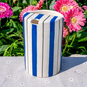 May include: A cylindrical ceramic vase featuring a white base with vertical blue stripes and thin black pinstripes. The top edge is curved, and there is a small, semi-circular cutout near the upper rim of the vessel, showcasing a smooth finish.