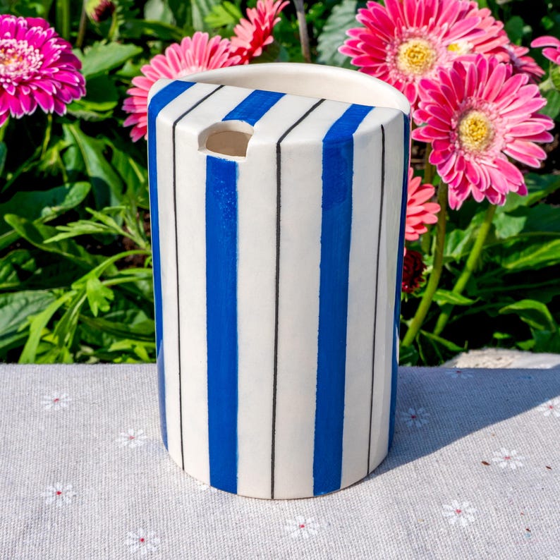 May include: A cylindrical ceramic vase featuring a white base with vertical blue stripes and thin black pinstripes. The top edge is curved, and there is a small, semi-circular cutout near the upper rim of the vessel, showcasing a smooth finish.