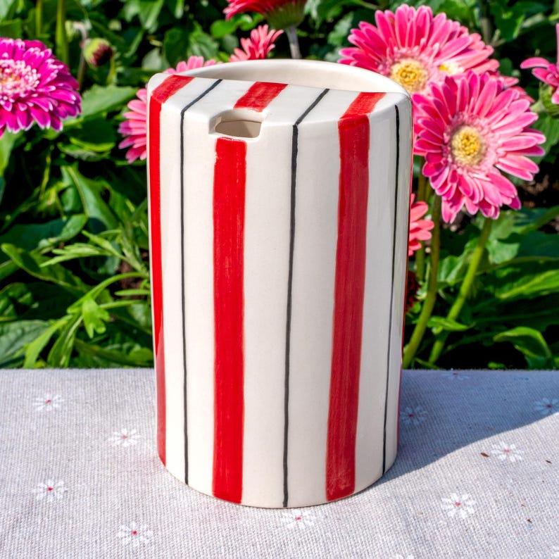 May include: A cylindrical ceramic utensil holder featuring a white base with vertical red stripes and thin black pinstripes. The top edge has a small semi-circular cutout, and the surface has a glossy finish, showcasing a clean, structured design.