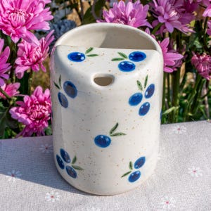 May include: A cylindrical, off-white ceramic wall pocket with a speckled finish. It features hand-painted blue olive branch designs. The pocket has a curved top and a hole for hanging. The background includes pink flowers.