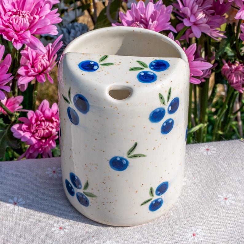 May include: A cylindrical, off-white ceramic wall pocket with a speckled finish. It features hand-painted blue olive branch designs. The pocket has a curved top and a hole for hanging. The background includes pink flowers.