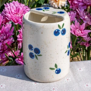 May include: A ceramic toothbrush holder with a cream-colored speckled finish. The holder is decorated with hand-painted blue blueberries and green leaves. The holder has a curved top with a slot for toothbrushes.